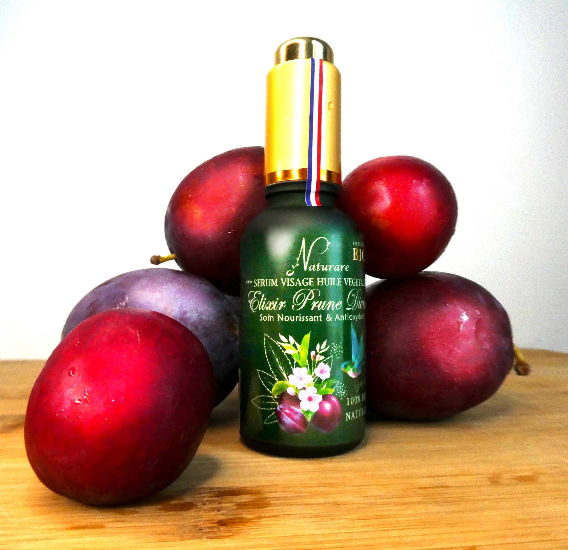 Agnès' Petit Paris Skincare organic plum facial serum bottle surrounded by ripe plums on wooden surface